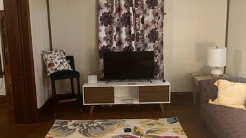 A living room with a flat-screen TV on a wooden TV stand, a floral-patterned rug, a purple sofa, and a small table with a lamp.