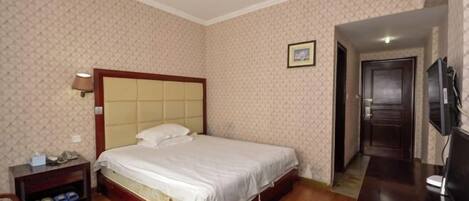A hotel room with a bed, a bedside table, a lamp, a TV, and a door leading to another room.