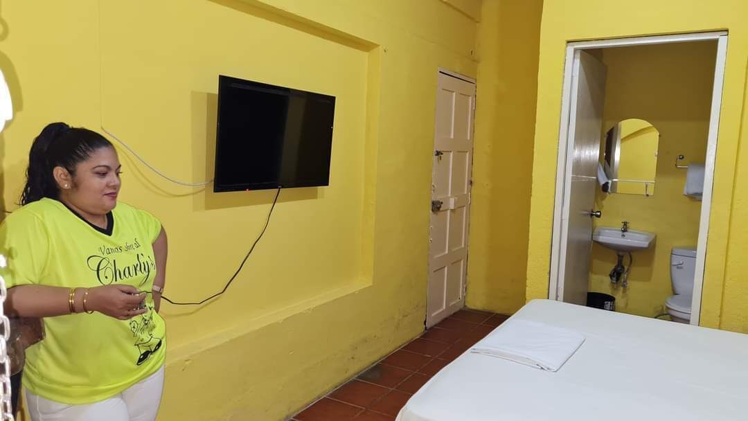 Una habitación con paredes amarillas, una cama con colchón blanco, un televisor montado en la pared y un baño visible a través de una puerta abierta.