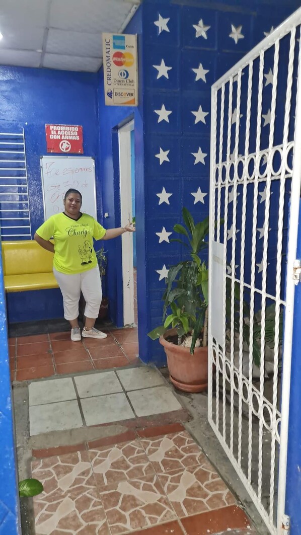 Una persona con camisa verde de pie frente a una puerta, con una pared azul y estrellas blancas, una pizarra y un letrero que prohíbe el acceso a personas con los brazos.