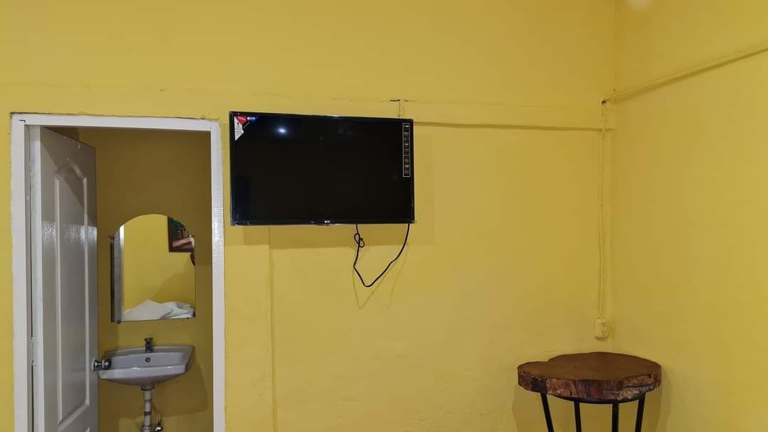 Une pièce avec un mur jaune, un téléviseur fixé au mur, un petit lavabo et une table ronde en bois.