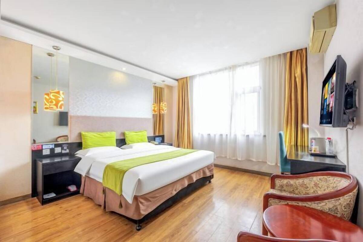 Habitación de hotel con cama, mesitas de noche, escritorio, silla, televisor y un ventanal grande con cortinas.