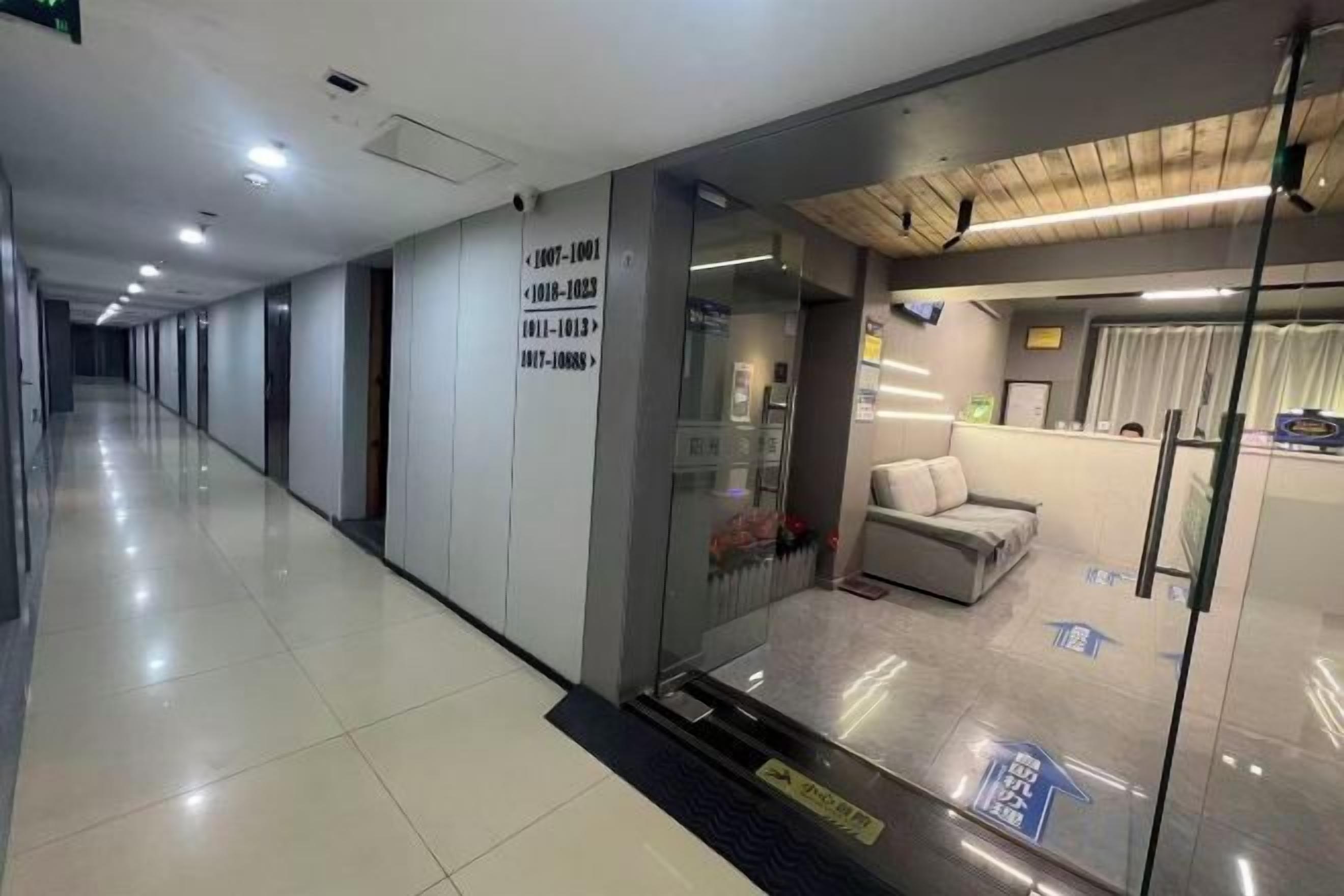 A hotel corridor with a reception area featuring a sofa, a glass partition, and a sign with room numbers.