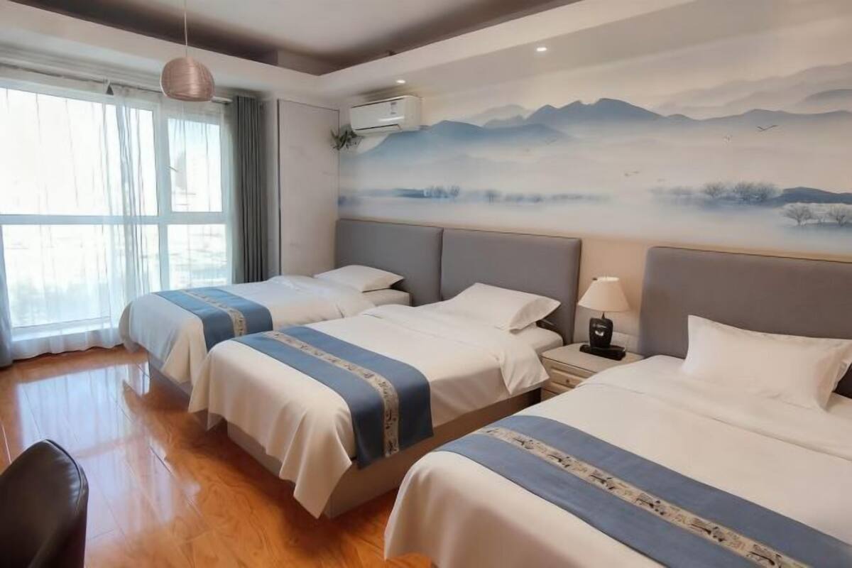 A hotel room with two beds, a large window with curtains, a wall-mounted air conditioner, and a painting of a landscape.