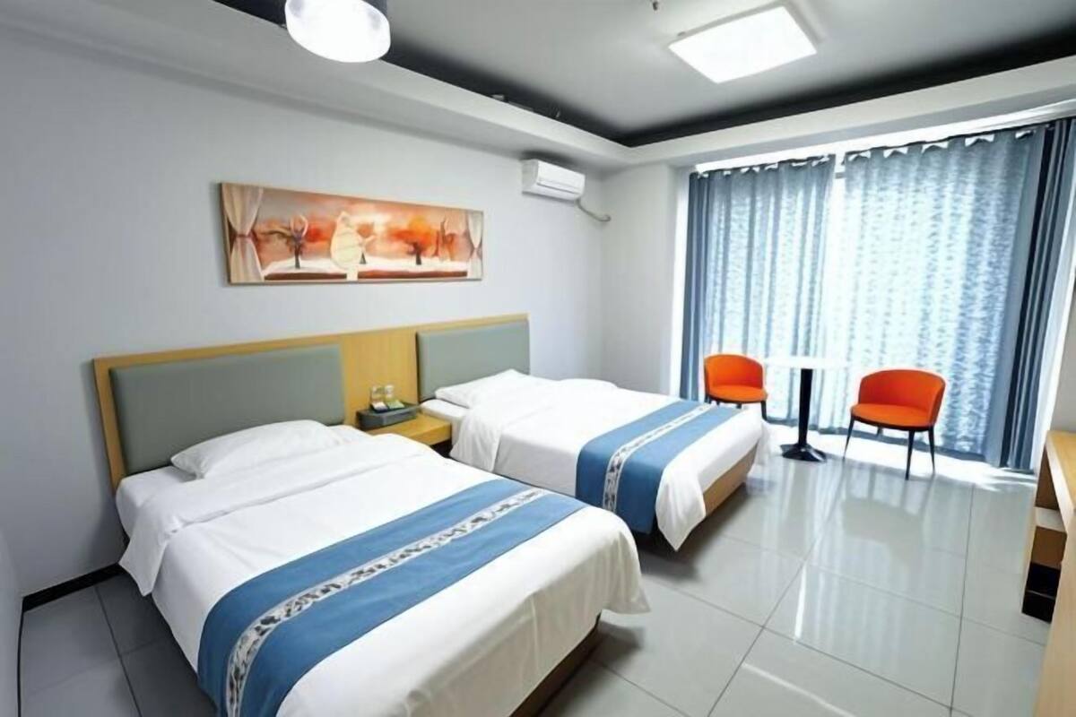 A hotel room with two beds, a painting on the wall, a window with curtains, and a small table with chairs.