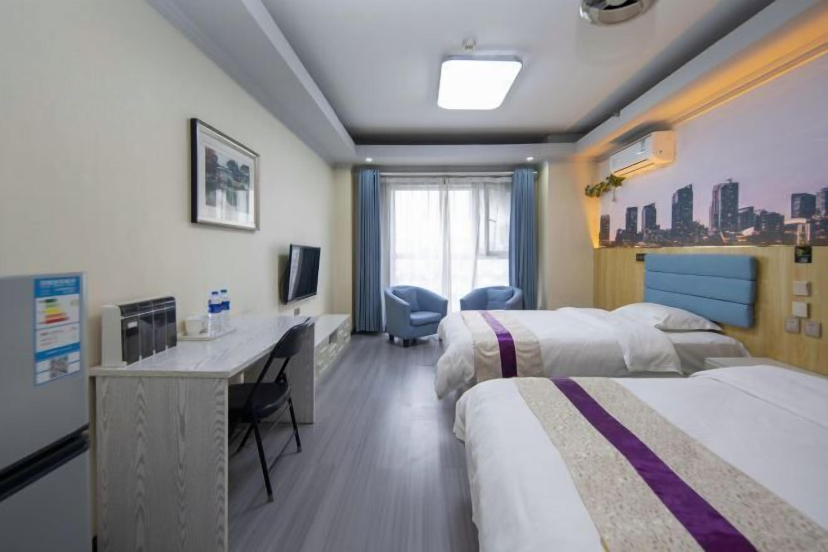 A hotel room with two beds, a desk with a chair, a TV, and a cityscape mural on the wall.