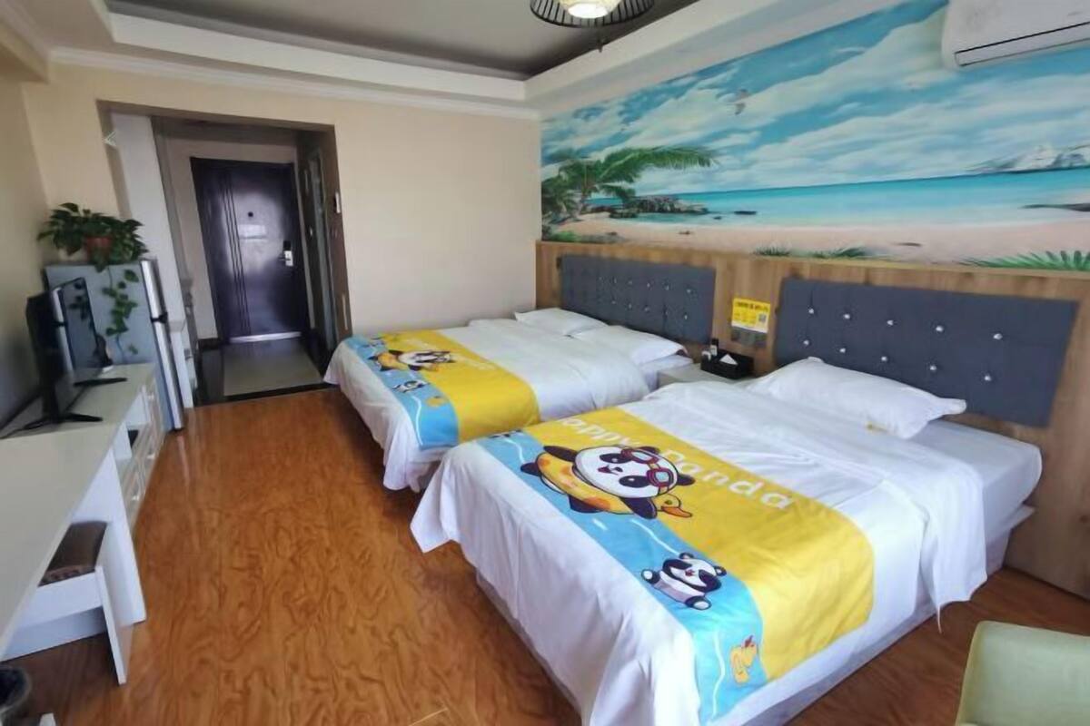 A hotel room with two beds, a painting of a beach scene, a refrigerator, and a desk.