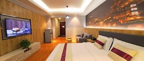 A modern hotel room with a large bed, a flat-screen TV mounted on the wall, a sofa, and a dining table.