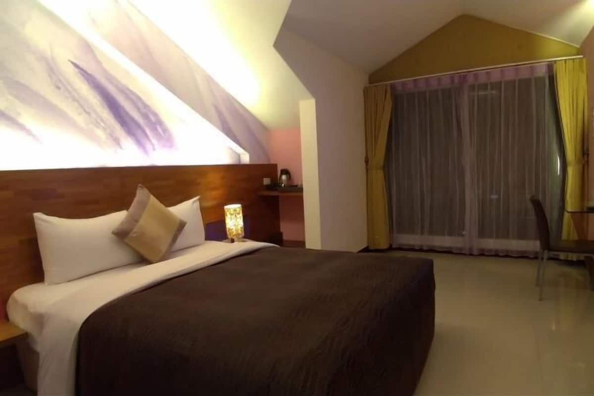 Hotellihuone, jossa on suuri sänky, yöpöytä, lamppu, tuoli ja ikkunasta avautuva näkymä ulos, ikkunassa on verhot.