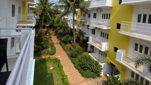 A courtyard with a paved walkway, surrounded by greenery and palm trees, with multi-story buildings featuring balconies and white and yellow exteriors. - VISTA Montanha  GOA (Calangute)