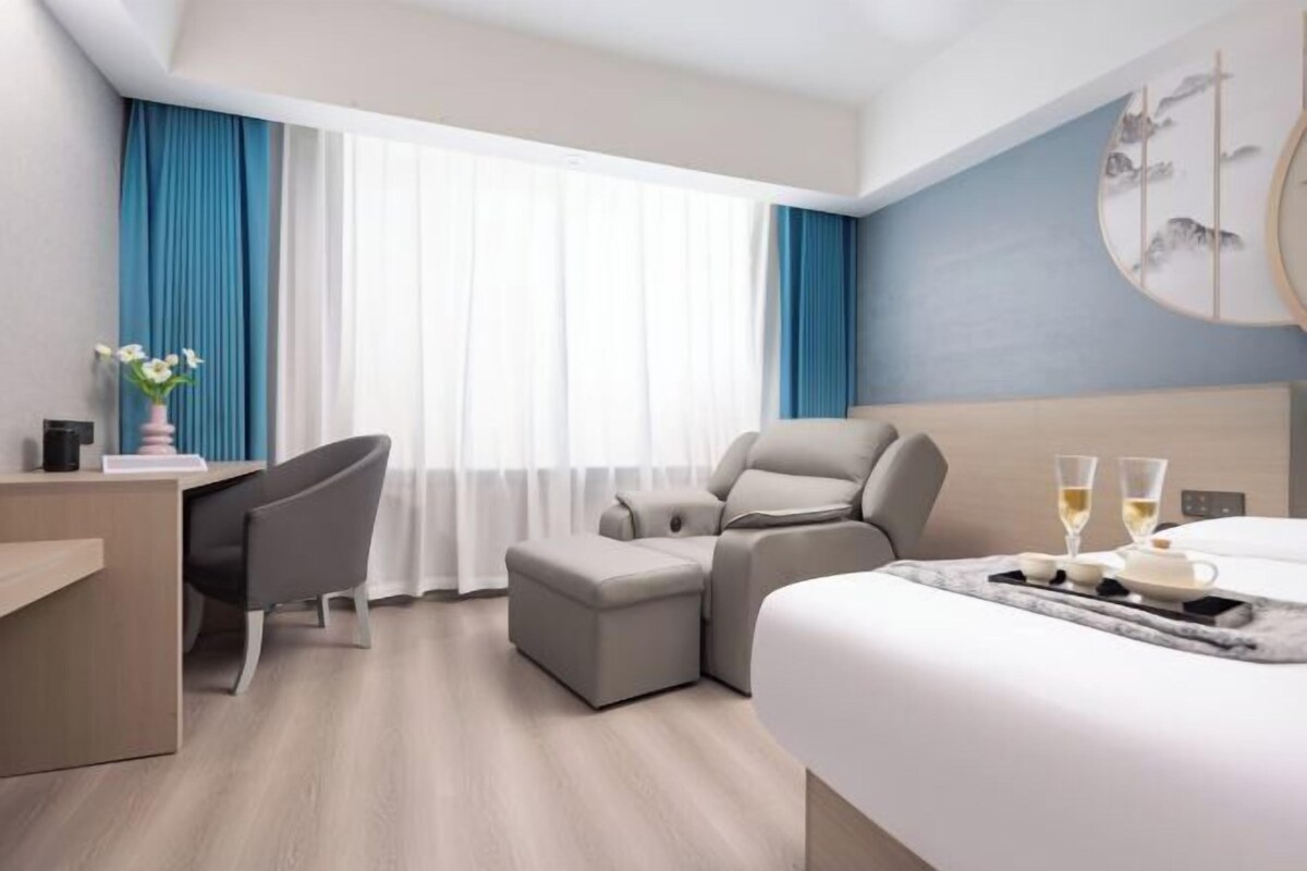 Ein modernes Hotelzimmer mit Bett, Schreibtisch, Sessel, einer Couch und einem großen Fenster mit Vorhängen.
