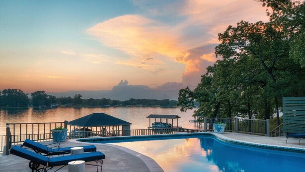 Ein Swimmingpool mit Liegestühlen, einer Laube und Blick auf einen See bei Sonnenuntergang.