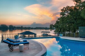 Piscina com cadeiras de descanso, um gazebo e vista para um lago ao pôr do sol.