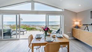 Un espace repas moderne avec vue sur la mer, une table dressée pour deux personnes et un télescope posé sur un trépied.