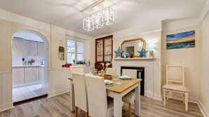 A dining area with a wooden table, white chairs, a chandelier, a fireplace, and a painting on the wall. - 2 Bed in Appledore (oc-s29561) (Appledore)
