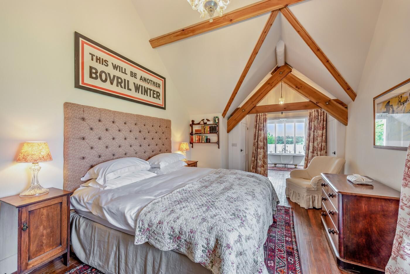 Een slaapkamer met een groot bed, houten balken, een boekenkast, een bureau en uitzicht op het omliggende landschap.
