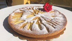 Ein runder Kuchen mit einer Zuckerstaubschicht und einer roten Blume, platziert auf einem weißen Teller auf einer Tischplatte.