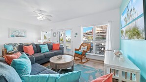 A living room with a grey sofa, teal and coral cushions, a wooden coffee table, a rocking chair, and a view of a building through large windows.