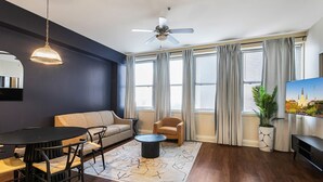Un salon moderne avec un mur d’accent bleu foncé, une table à manger ronde avec des chaises, un canapé, un ventilateur de plafond et une grande fenêtre avec des rideaux.