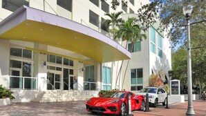 Un hotel moderno con un toldo de entrada curvo, un coche deportivo rojo estacionado al frente y un coche blanco cerca.