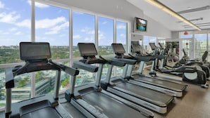 A gym with treadmills and exercise equipment, large windows with a city view, and a television mounted on the wall.