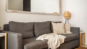 Ein graues Sofa mit weißer Decke und zwei gemusterten Kissen, ein großer Spiegel an der Wand und eine gewebte Stehlampe.