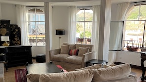A spacious living room with a large beige sofa, a black coffee table, and a floor lamp. There is a window with a view of a balcony and buildings.