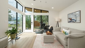 Un salon moderne avec un canapé gris, une table basse, un fauteuil en cuir noir, un ventilateur de plafond et une grande fenêtre donnant sur l’extérieur.