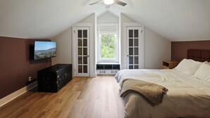 A bedroom with a sloped ceiling fan, a large bed with a blanket, a television on the wall, and a window with a view of greenery.