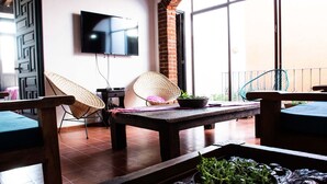 Un salon confortable avec une table en bois, deux fauteuils en osier, un téléviseur à écran plat fixé au mur et un balcon doté d’une balustrade.