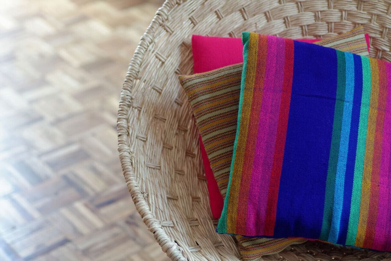 A wicker basket containing a colorful striped pillow and a pink fabric.