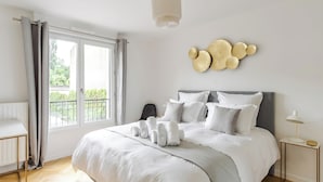 A modern bedroom with a large bed, a bedside table, a lamp, and a view of the outdoors through a window. - Appart. Victoria(sleeps 7, 2 parking spaces)- Les Demoiselles à Versailles (Versailles)