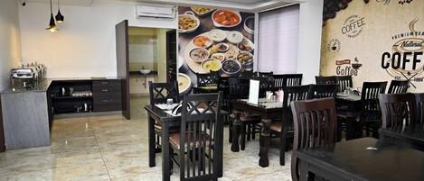 Un comedor con varias mesas y sillas, una cocina con estufa y armarios, y una pared con un mural alusivo al café.