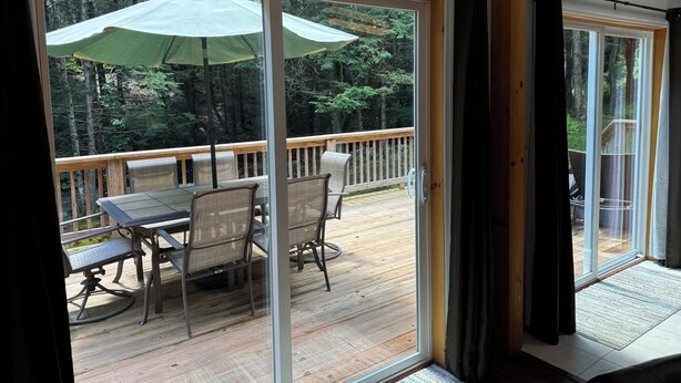 A deck with a table and chairs, a wooden railing, and a view of a forest through glass doors. - Waterfall View from Secluded 4 Bed Cabin in East Branch, NY with Mountain Views (East Branch)