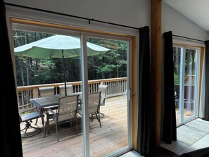 En uteplats med ett bord och stolar, ett träräcke och utsikt över en skog genom glasdörrar.
