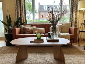 Ein gemütliches Wohnzimmer mit einem runden Couchtisch, einem braunen Sofa und einem großen Fenster mit Vorhängen.