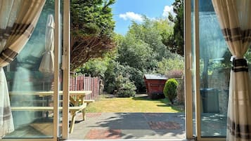A room with a sliding glass door leading to a backyard with a wooden table and chairs, a small shed, and a well-maintained lawn.
