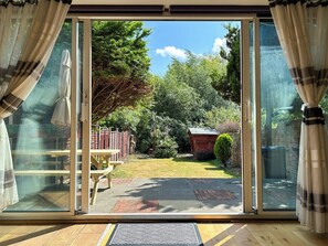 Une pièce dotée d’une porte-fenêtre coulissante donnant sur un jardin avec une table et des chaises en bois, un petit abri de jardin et une pelouse bien entretenue.
