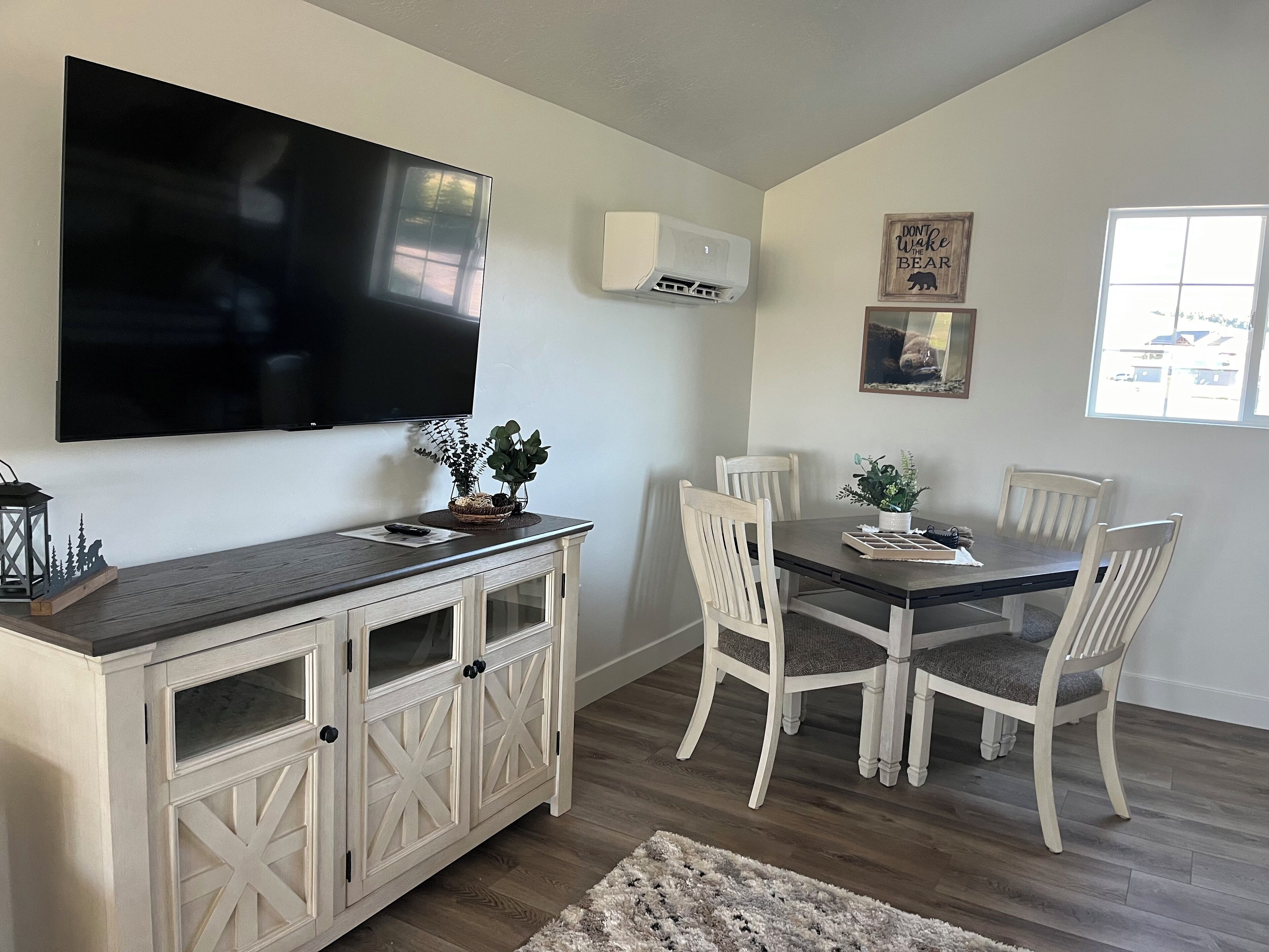 A dining area with a TV mounted on the wall, a wooden cabinet with glass doors, and a dining table with four chairs.