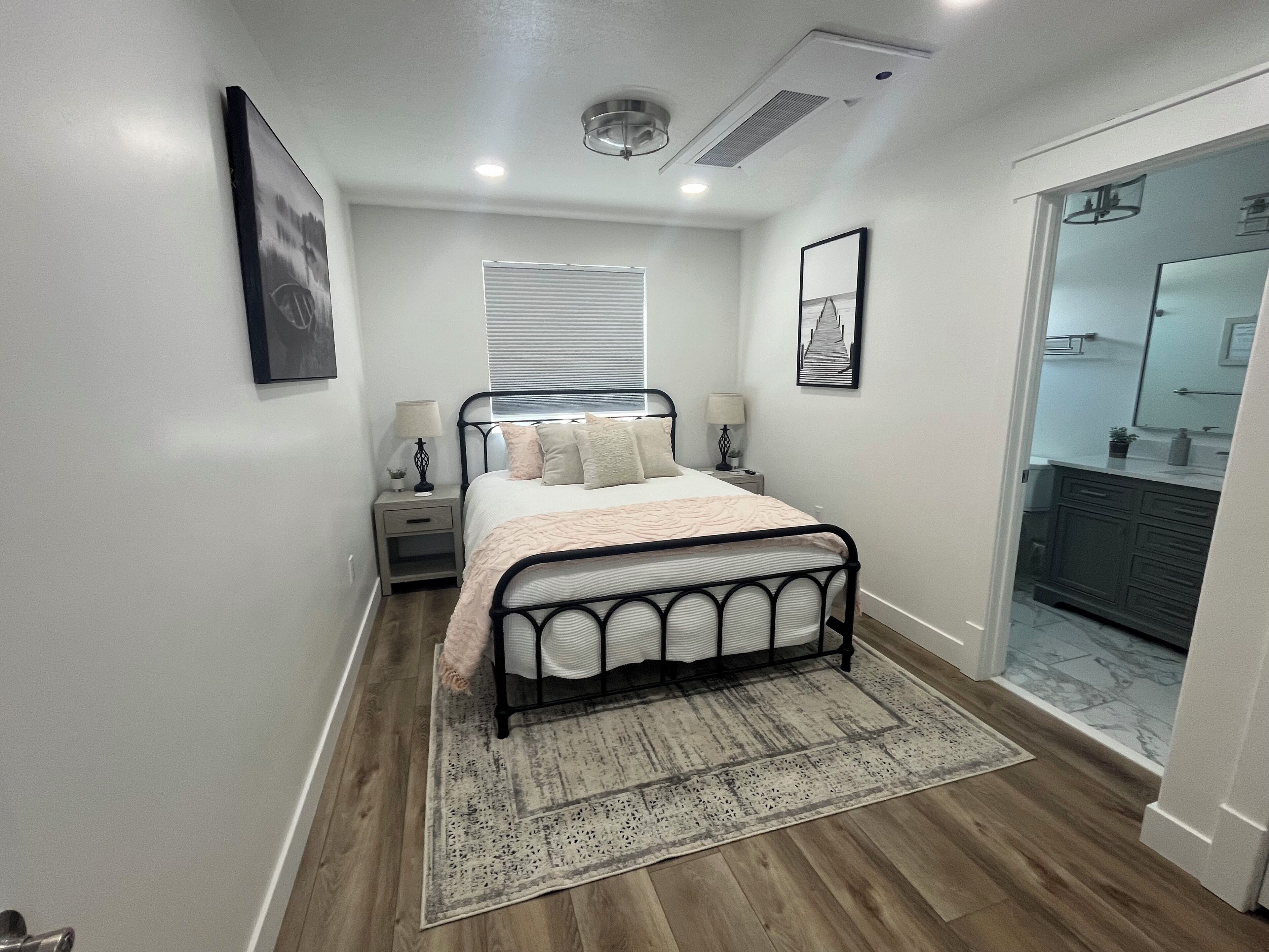 A bedroom with a bed, bedside tables, a nightstand, a window with blinds, a ceiling fan, and a bathroom visible through an open door.