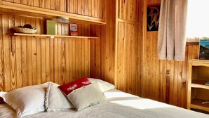 Een houten kamer met een bed, kussens, een plank met spullen en een raam met gordijnen.