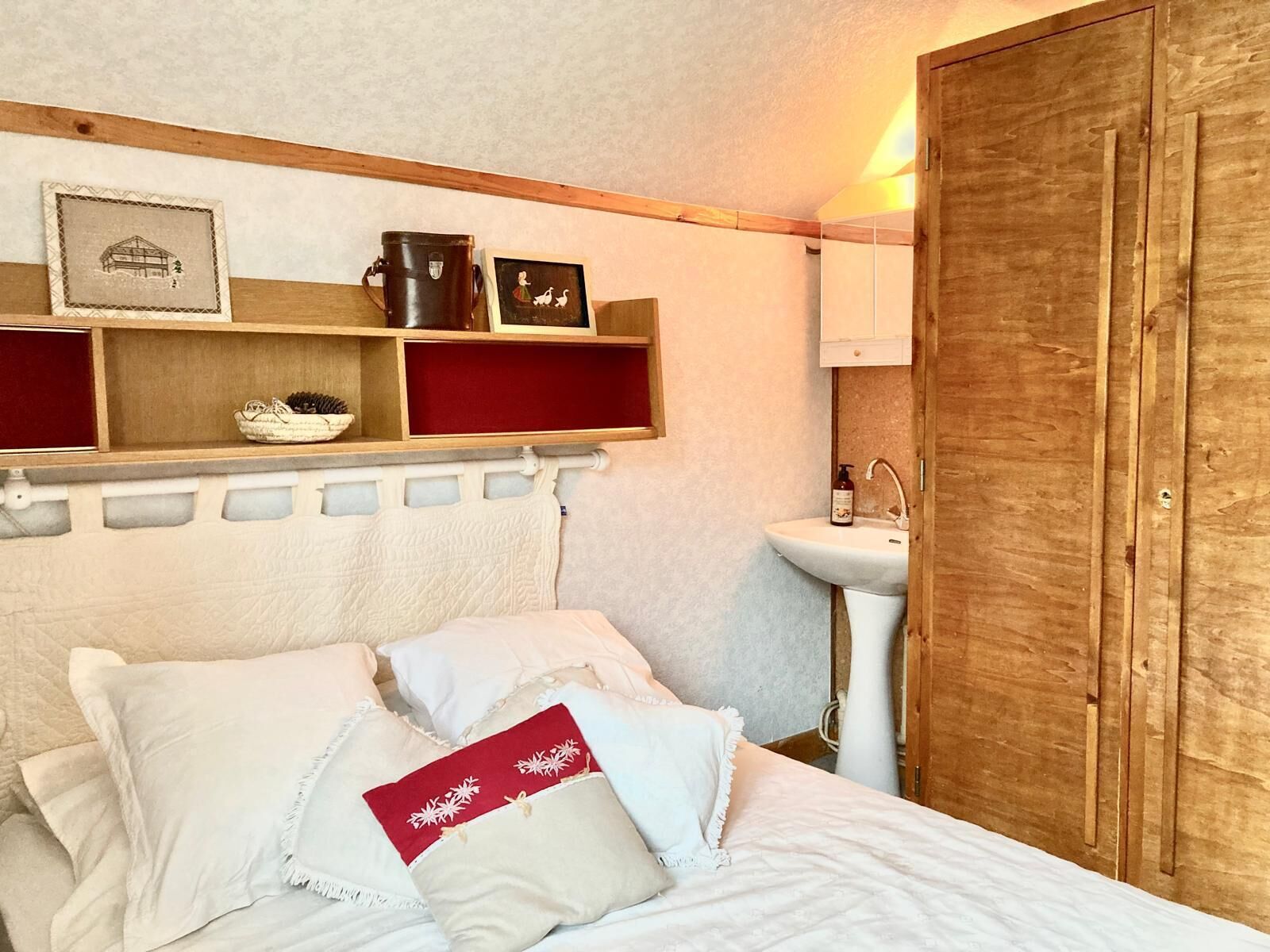 Een slaapkamer met een bed, kussens, een plank met decoratieve spullen, een houten garderobe en een witte wastafel.