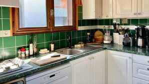 Een keuken met een gasfornuis, spoelbak en diverse apparaten. Er zijn kastjes, een raam en een groene betegelde achterwand.