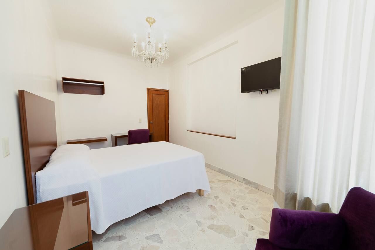 Habitación de hotel con cama, escritorio, silla, televisor y candelabro.