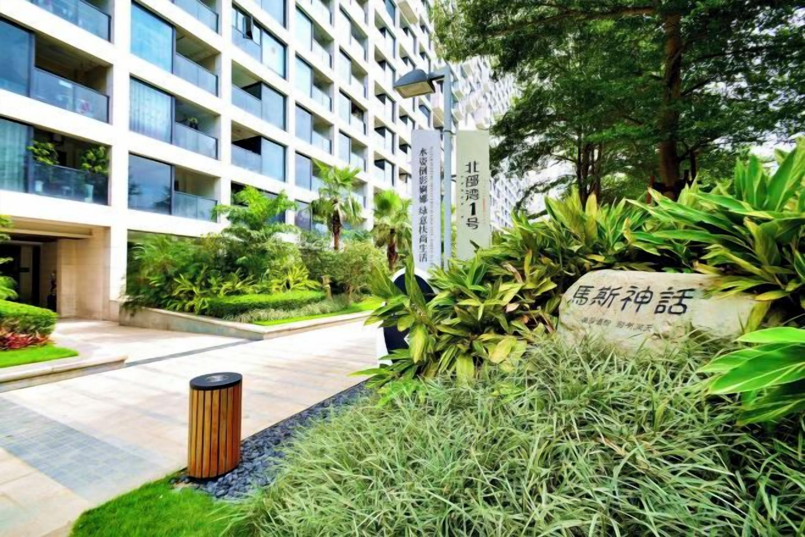 A modern building with a well-maintained garden area, a sign with Chinese characters, and a trash bin.