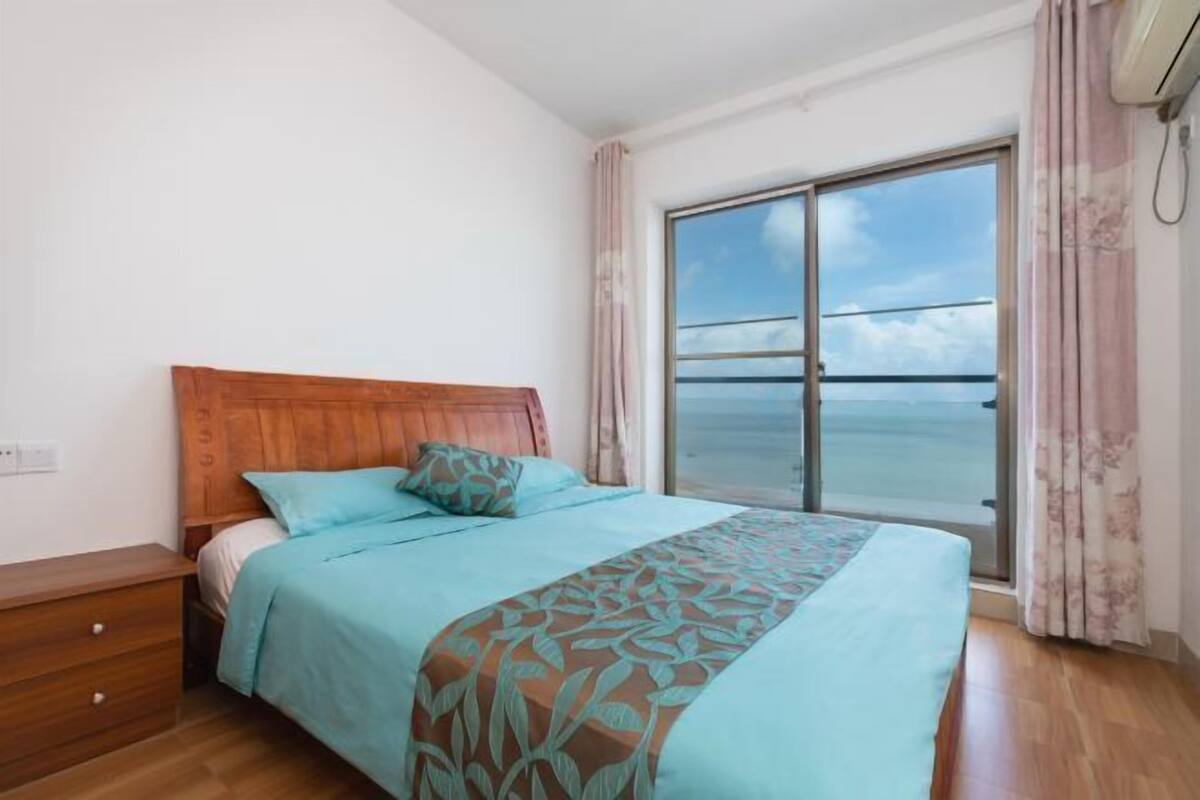 A bedroom with a wooden headboard, a bed with a patterned bedspread, a nightstand, and a large window with a view of the sea.
