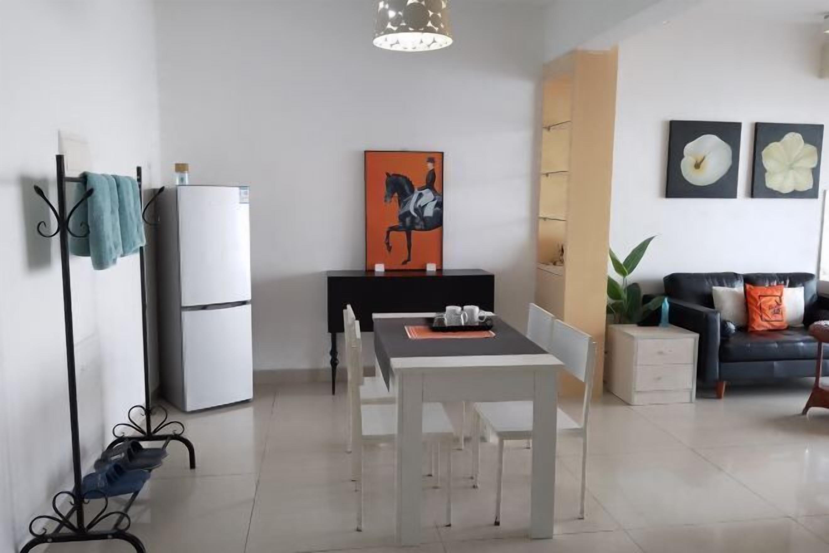 A modern living room with a dining area, a black sofa with orange cushions, a white refrigerator, and a painting of a horse.