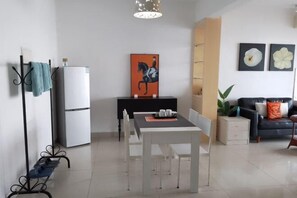 Un salon moderne comprenant un coin repas, un canapé noir avec des coussins orange, un réfrigérateur blanc et un tableau représentant un cheval.