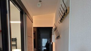 Un pasillo estrecho con piso de madera que conduce a una habitación oscura. Un perchero con ganchos está fijado a la pared.