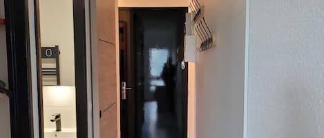 A narrow hallway with a wooden floor leading to a dark room. A coat rack with hooks is mounted on the wall.